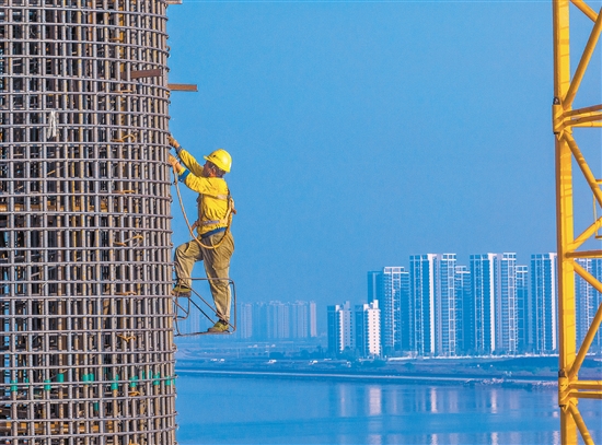 奮戰(zhàn)在百米索塔之上的建設者身影，與遠處的高樓大廈遙呼相應。